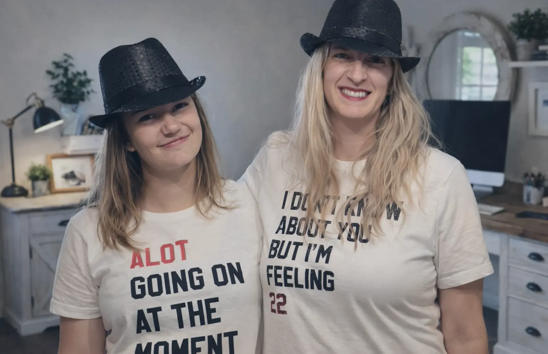 Mother and daughter web developer team stand in an office together. They are wearing t-shirts with Taylor Swift quotes saying "alot going on at the moment," and "I don't know about you but I'm feeling 22."