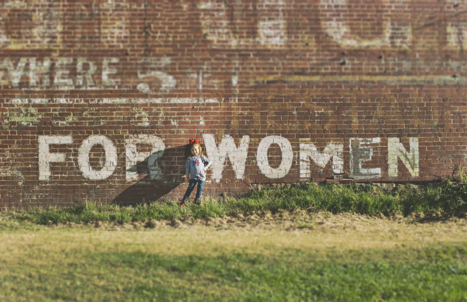 A young girl standing in grass, in front of a brick wall with writing on the wall hat says "for women."