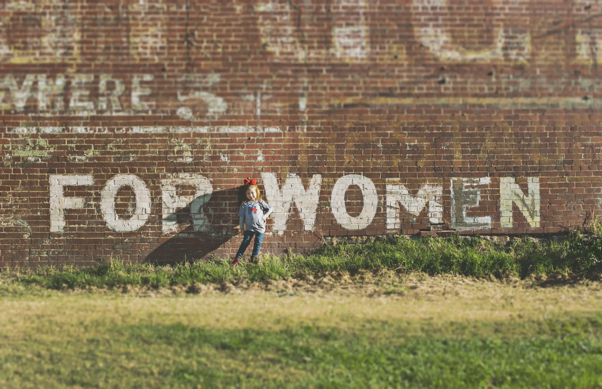 A young girl standing in grass, in front of a brick wall with writing on the wall hat says "for women."