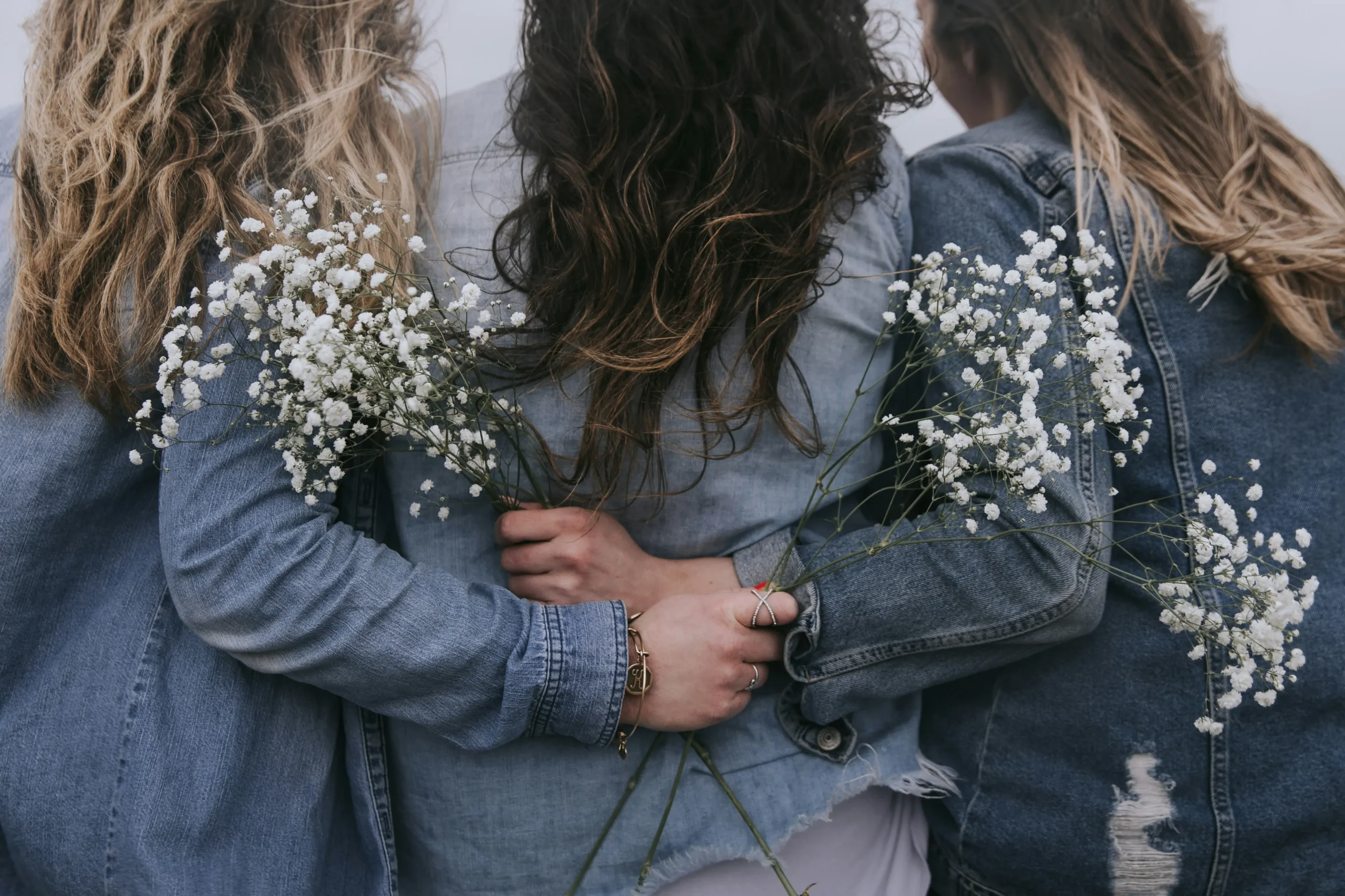 Three women stand together unified as authors, coaches, and course creators who emower other women. They wear denim jackets and hold delicate flowers symbolizing the strength and softness of women.