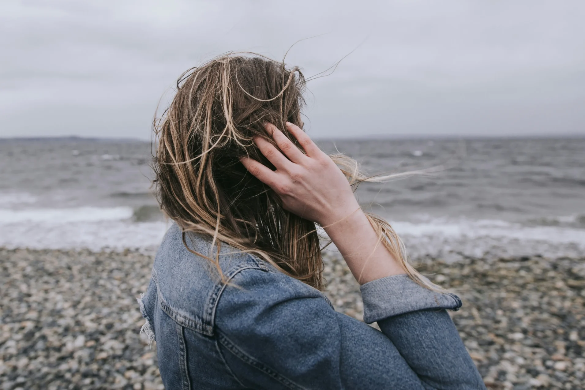 Woman wearing denim jacket stands alone and lost by the ocean on a rocky seaside.