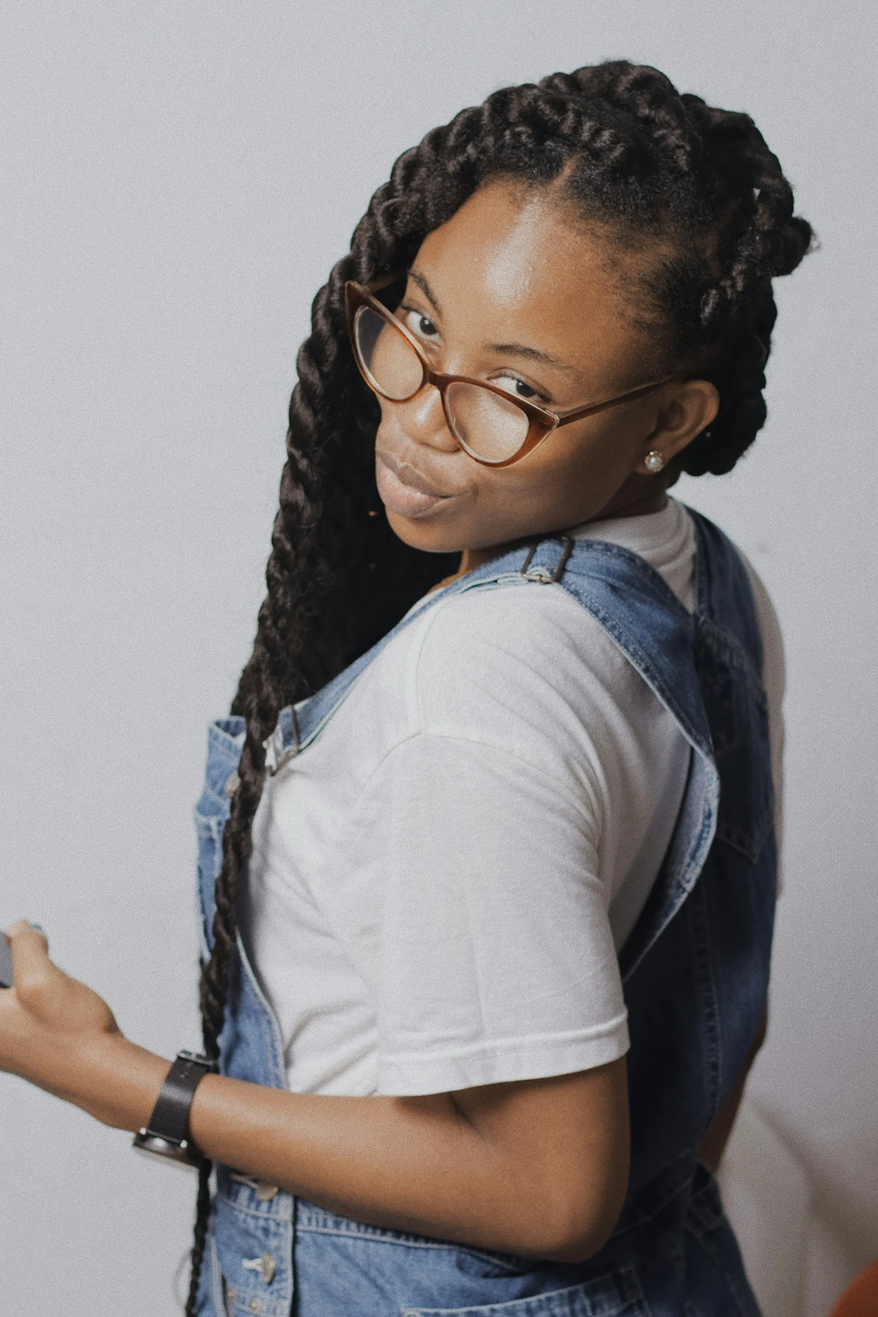 A woman with long black hair, wearing glasses and dressed in denim overalls and a white t-shirt turns and looks back at the camera.