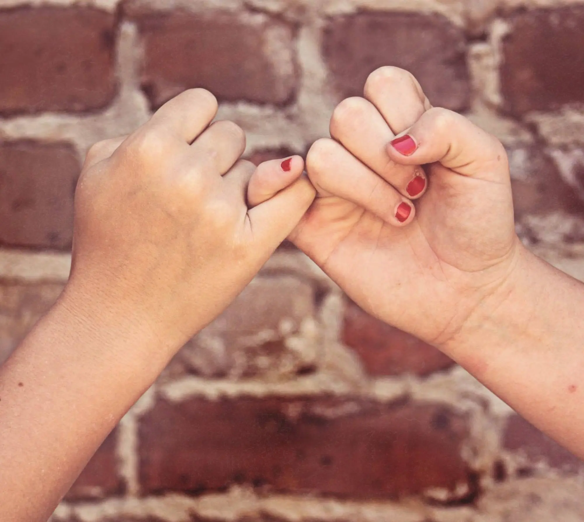 Two sister's hands in a pinky promise in front of a brick wall.