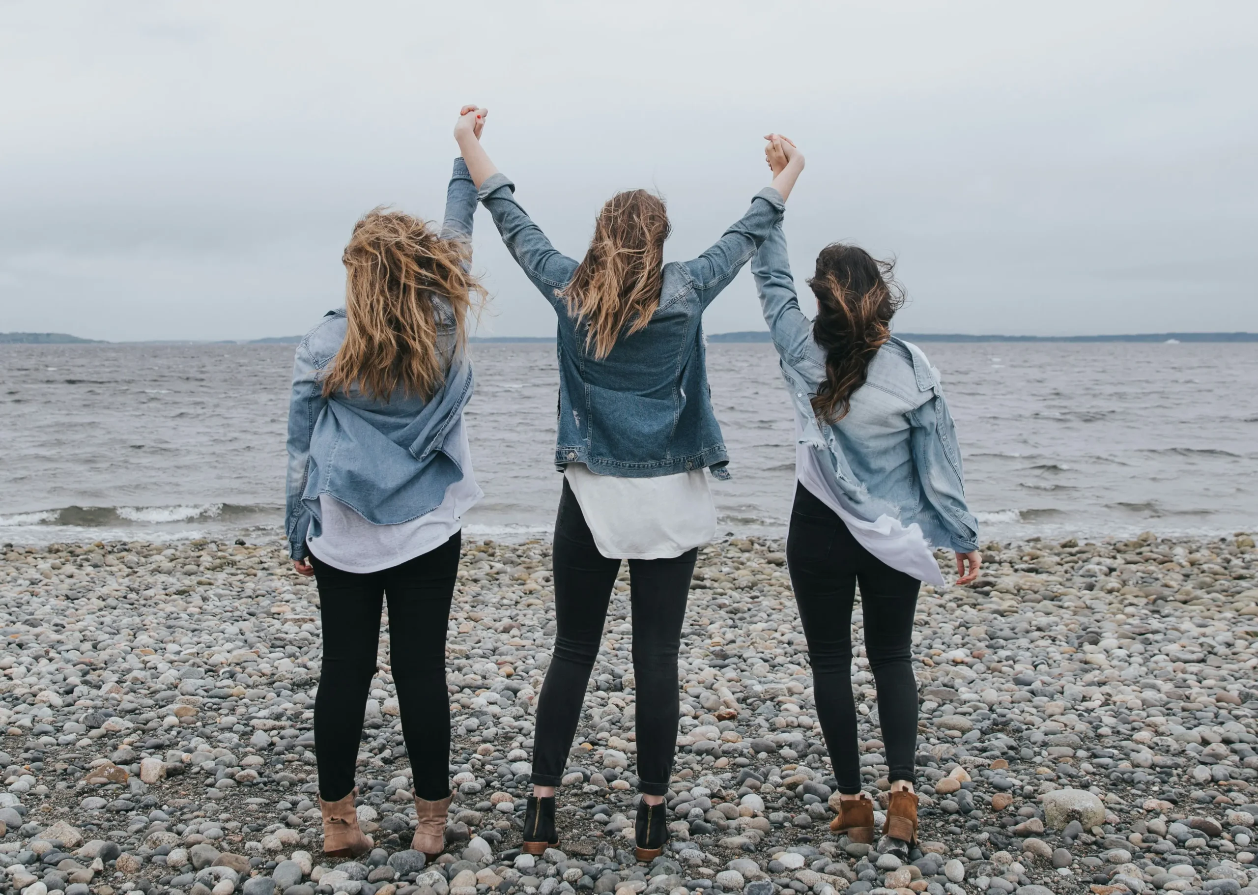 Portfolio Three women wearing denim jackets and jeans stand on a beach in celebration of empowerment with their hands locked together and held high.